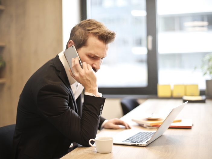 Business man sits in front of his laptop and takes a call from his cell phone that's clearly frustrating him.